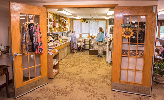 View through open double wooden doors into a small shop area inside a senior living facility. The shop has shelves stocked with various items and three elderly women are present, two standing and one sitting. The room is well-lit with fluorescent lights and has a carpeted floor.