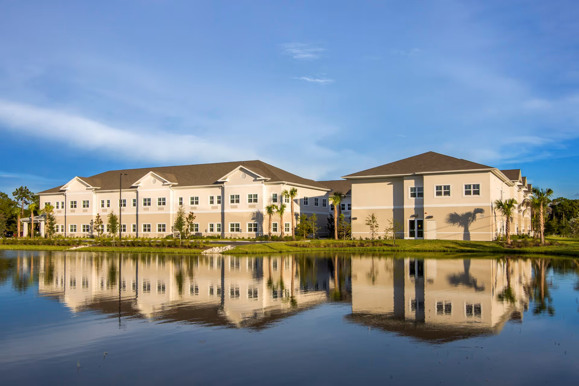 A large two-story beige and white building reflected in a calm pond under a blue sky with some clouds. The building is surrounded by grass, small trees, and palm trees.