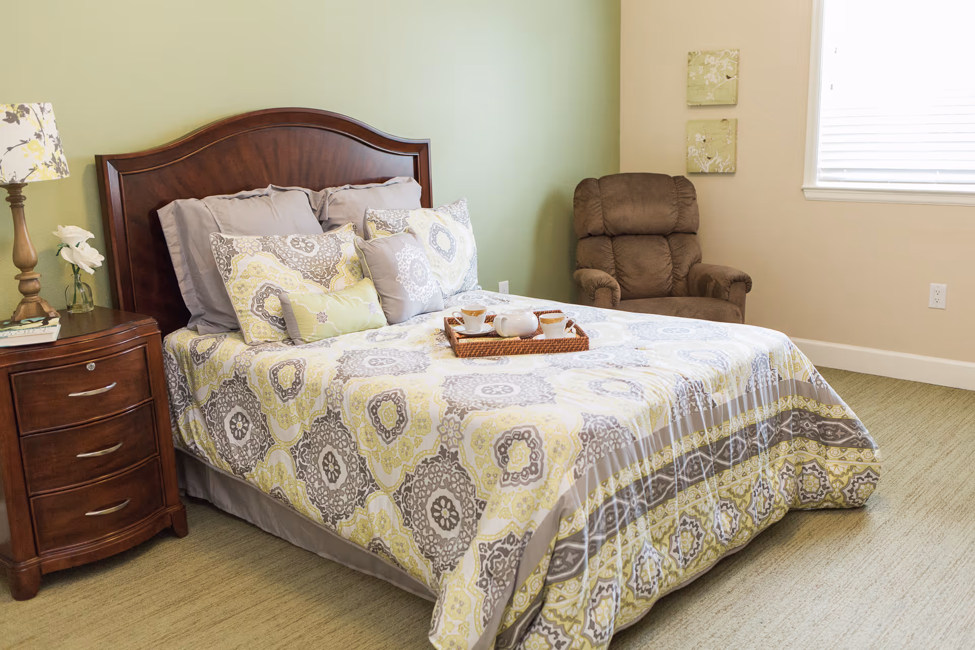 A neatly made bed with a patterned yellow, gray, and white bedspread and multiple pillows in a bedroom. There is a wooden nightstand with a lamp and a small vase with flowers on the left side of the bed. A brown cushioned armchair is positioned near a window with blinds, and two small decorative wall hangings are on the wall above the chair.