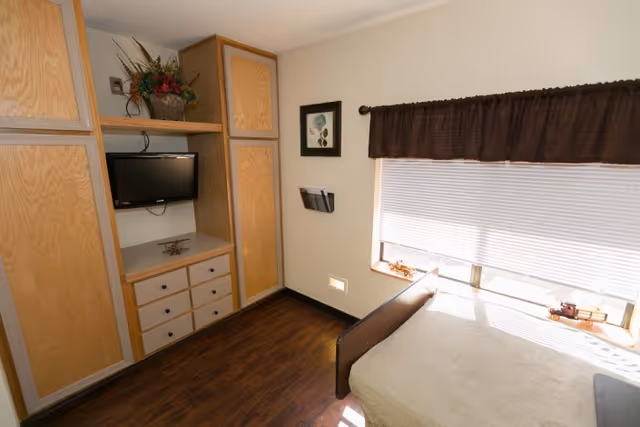 A small bedroom with wooden flooring, a bed covered with a light-colored blanket near a window with closed blinds and a dark valance. There is a built-in wooden cabinet with drawers and a mounted flat-screen TV. A framed picture and a wall-mounted magazine holder are on the wall next to the window.