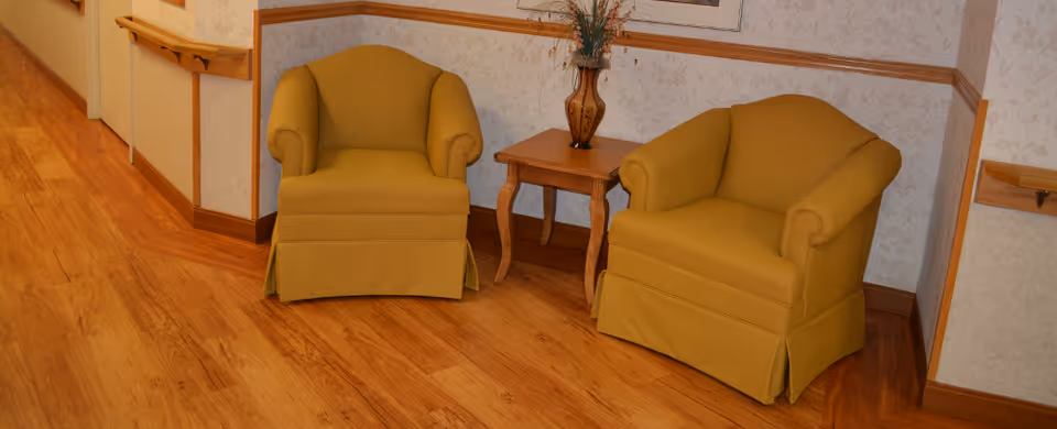 Two mustard yellow upholstered armchairs placed side by side in a corner with a small wooden table between them holding a decorative vase with flowers. The room has wooden flooring, light-colored wallpaper with a subtle pattern, and wooden chair rails along the walls.