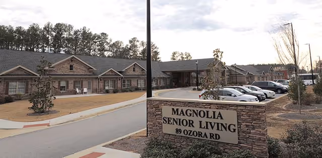Exterior view of Magnolia Senior Living at Sugar Hill, showing a single-story brick building with a parking lot in front and a stone sign displaying the facility's name and address, 89 Ozora Rd.
