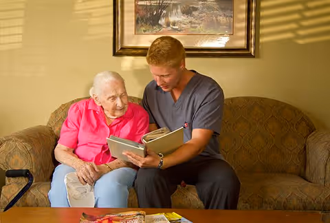 An elderly woman in a pink shirt sitting on a patterned couch next to a caregiver in gray scrubs who is showing her a photo album. They are in a warmly lit room with a framed painting on the wall behind them and a wooden table with magazines in front of them.