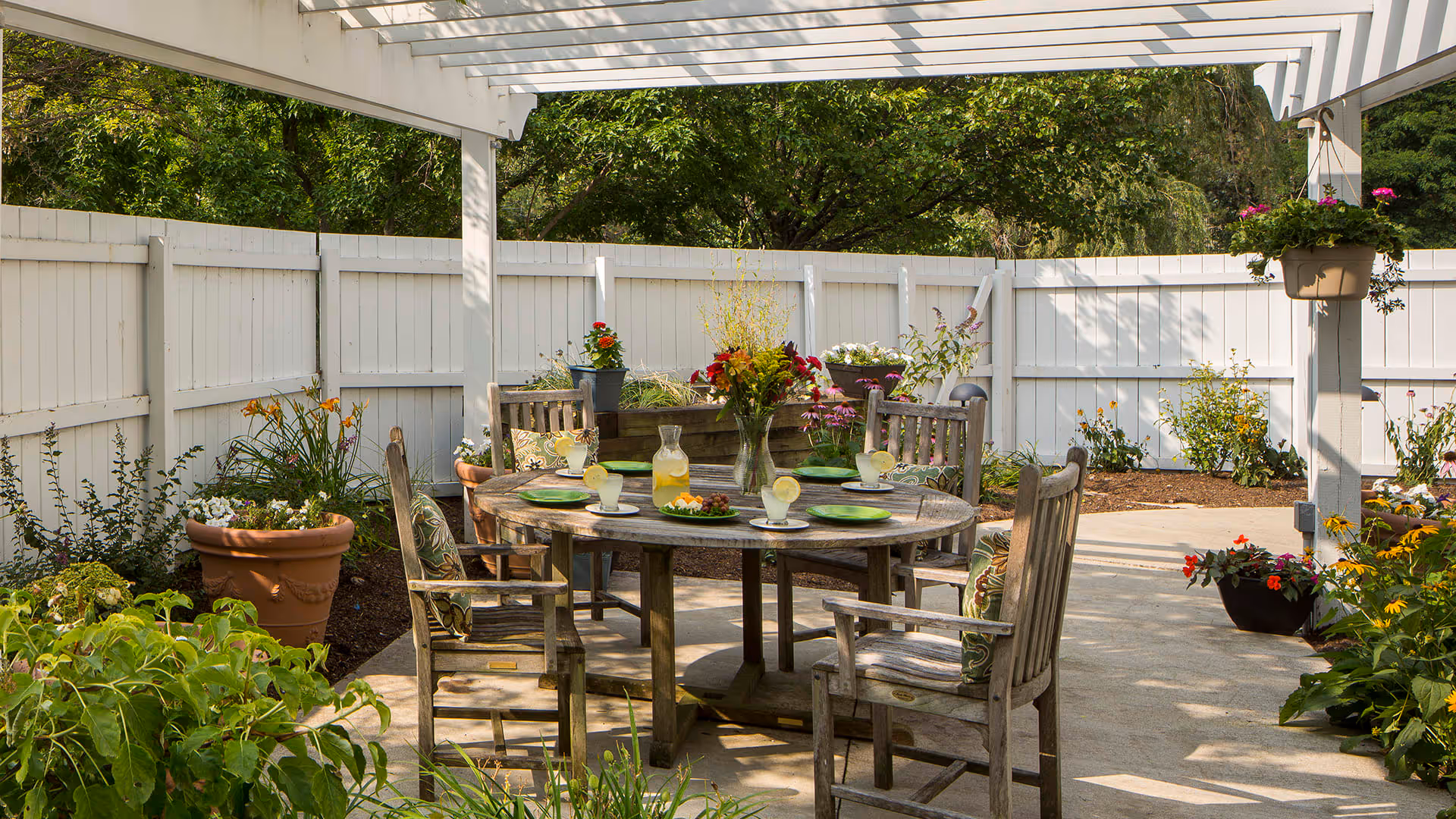 Outdoor patio area with a round wooden table set for four with green plates, glasses of lemonade, and a vase of flowers. The patio is surrounded by a white wooden fence and various potted plants and flowers. A pergola provides partial shade overhead.
