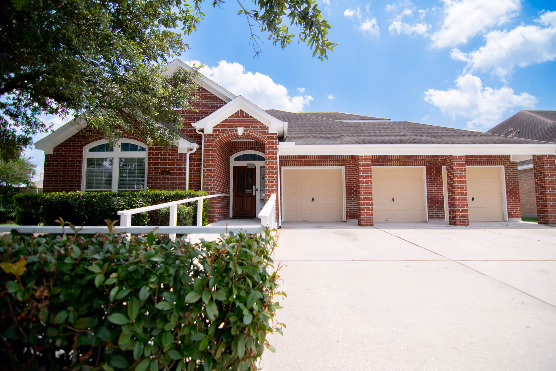 Exterior view of a brick building with a driveway leading to three beige garage doors. The building has a white-trimmed roof and a covered entrance with a wooden door. Green bushes and a tree are visible in the foreground under a blue sky with scattered clouds.