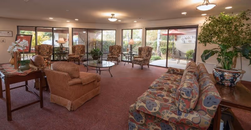 A cozy senior living common area with patterned armchairs and a sofa arranged around a glass coffee table. Large windows and sliding glass doors provide a view of an outdoor patio with chairs and greenery. The room is warmly lit with ceiling lights and table lamps, and decorated with plants and flowers.