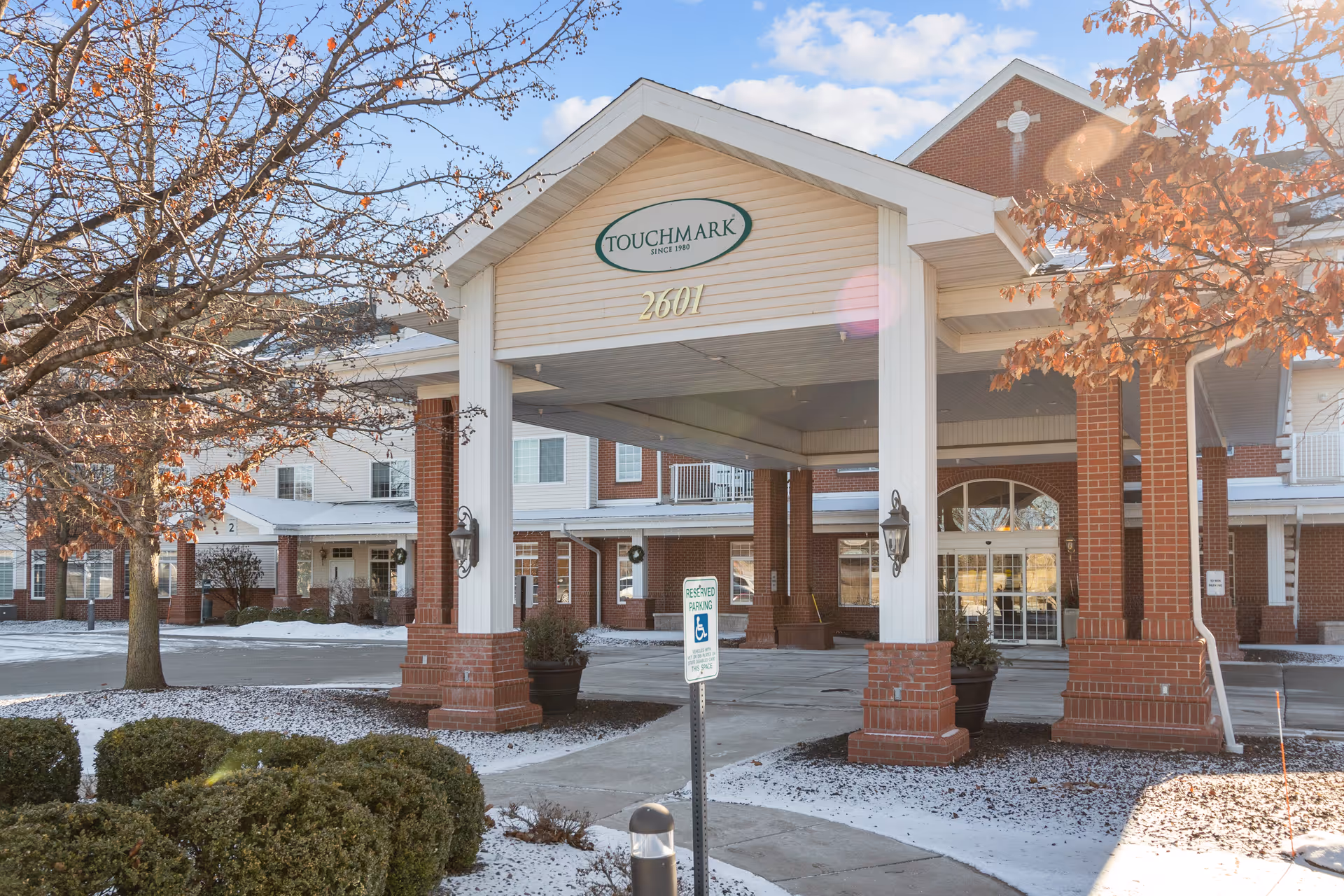 Covered main entrance and brick facade of the Touchmark on West Prospect building with light snow on the ground and nearby trees.