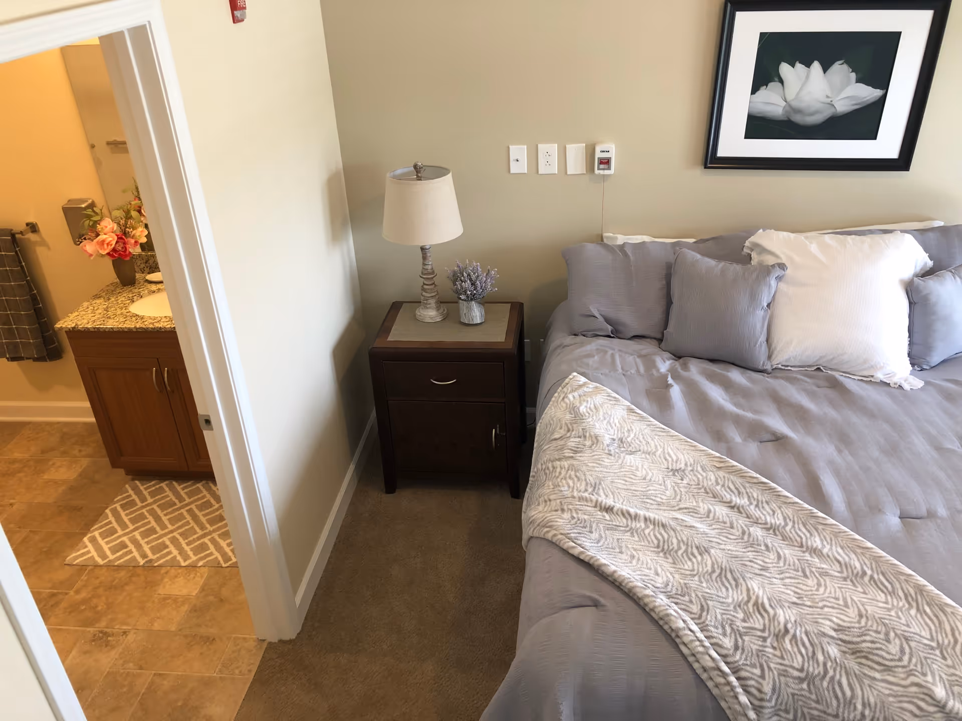 A bedroom with a made bed in gray bedding and pillows, a wooden bedside table with a lamp and small plant, and an open doorway showing a bathroom vanity.