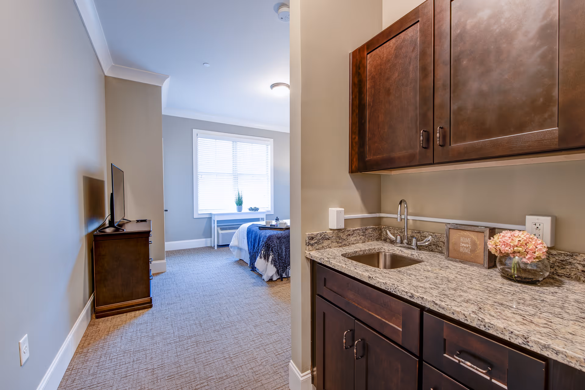 Interior view of a senior living facility room at Northshore Heights showing a small kitchenette area with dark wood cabinets, a granite countertop, a sink, a small decorative flower vase, and a framed sign that reads 'Home Sweet Home'. In the background, there is a bedroom area with a bed covered in white and blue bedding, a window with blinds, a small table with a plant, and a TV on a wooden dresser.