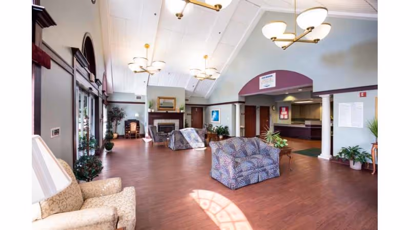 A spacious and well-lit common area in a senior living facility with high vaulted ceilings, multiple chandeliers, and large windows letting in natural light. The room features patterned upholstered armchairs and sofas arranged around a central fireplace. There are plants and framed artwork on the walls, with a reception or service counter visible in the background.