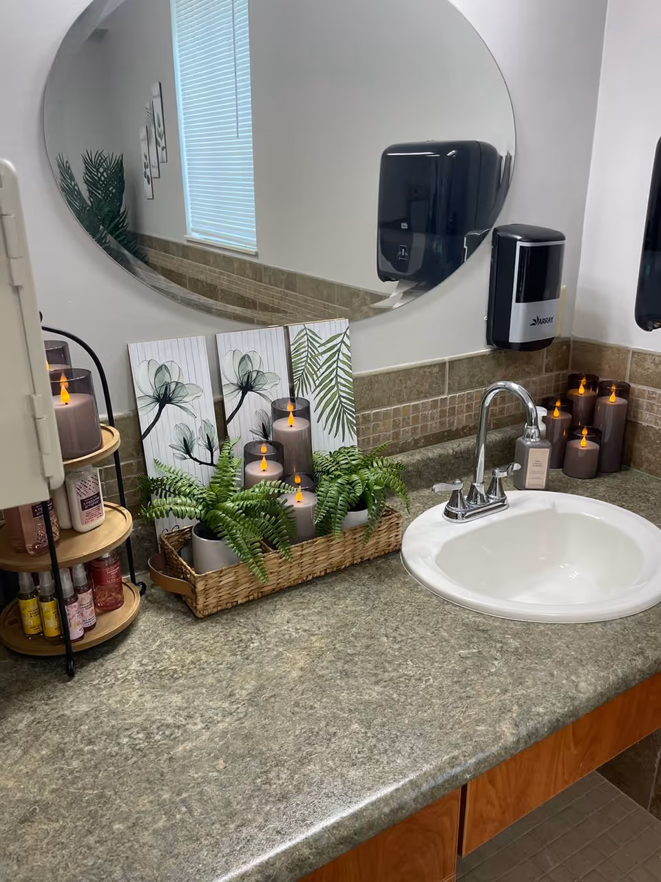 A bathroom countertop with a white sink and silver faucet. On the counter are decorative items including a basket with green ferns and several flameless candles, three floral and leaf artwork panels, and a small rotating shelf holding various bottles. Above the sink is a large round mirror, and mounted on the wall are a black paper towel dispenser and a black soap dispenser.