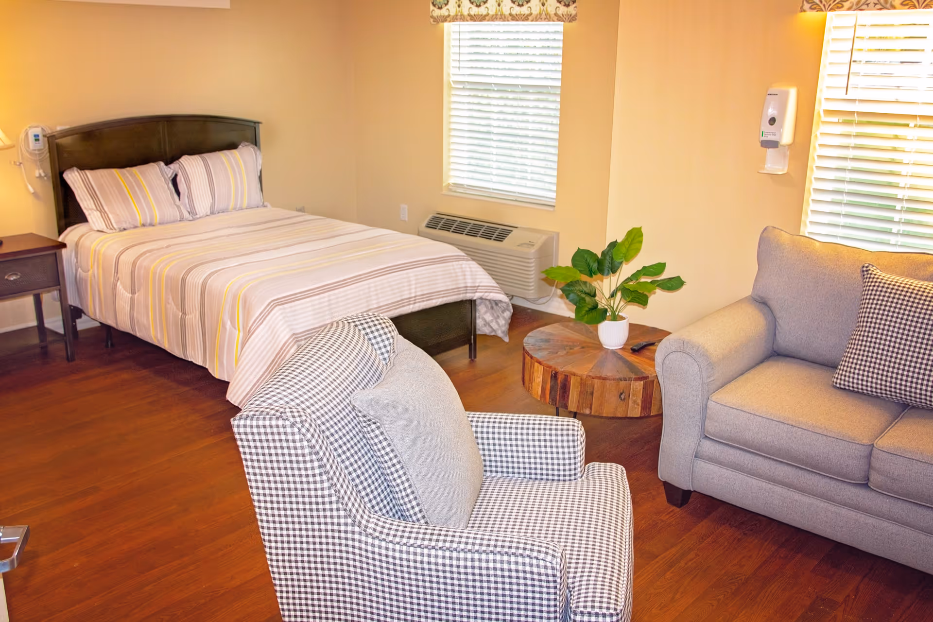 A cozy room with a bed featuring striped bedding, a wooden nightstand with a lamp, a checkered armchair, a gray sofa with a checkered pillow, a round wooden coffee table with a potted plant, and two windows with blinds. The room has wooden flooring and beige walls.