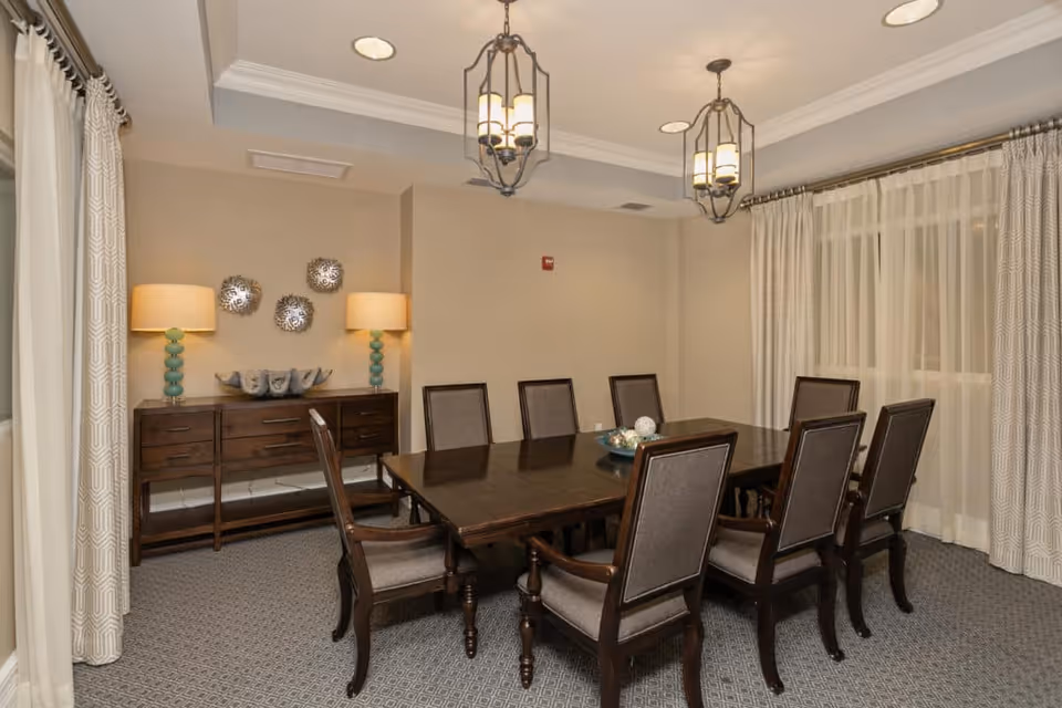 A formal dining room with a large dark wooden table surrounded by eight upholstered chairs. The room features beige walls, patterned carpet, two hanging pendant lights, and a sideboard with two lamps and decorative items. Large windows are covered with sheer and patterned curtains.