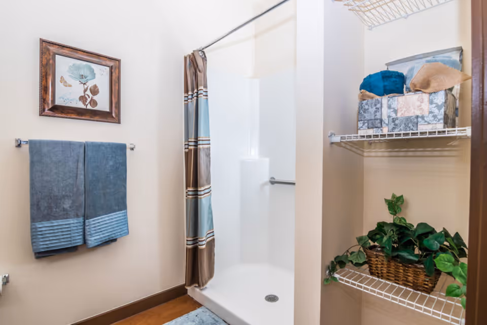 A bathroom with a white shower stall featuring a striped shower curtain. On the left wall, there is a framed floral picture above two blue towels hanging on a towel rack. On the right, there are two wire shelves holding a basket with green leafy plants and two fabric storage boxes with towels.
