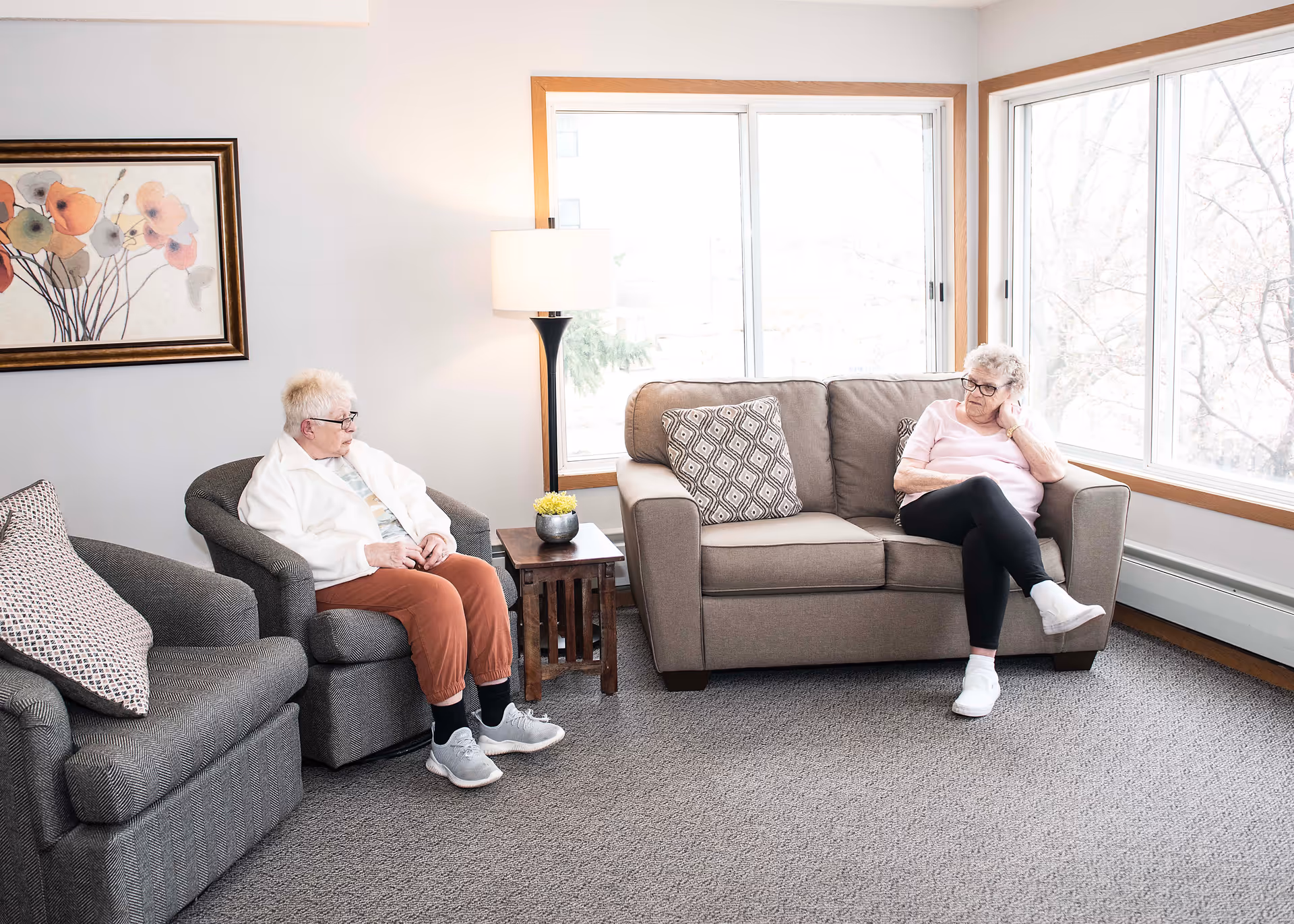 Two elderly women sitting in a bright living room with large windows. One woman is seated on a gray armchair wearing a white jacket and rust-colored pants, while the other woman is sitting on a beige sofa wearing a pink top and black pants. The room has a floor lamp, a small wooden side table with a small plant, and a framed floral painting on the wall.