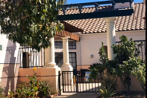 Gated entrance to a residential care building with white columns, a tiled roof pergola, and surrounding plants.