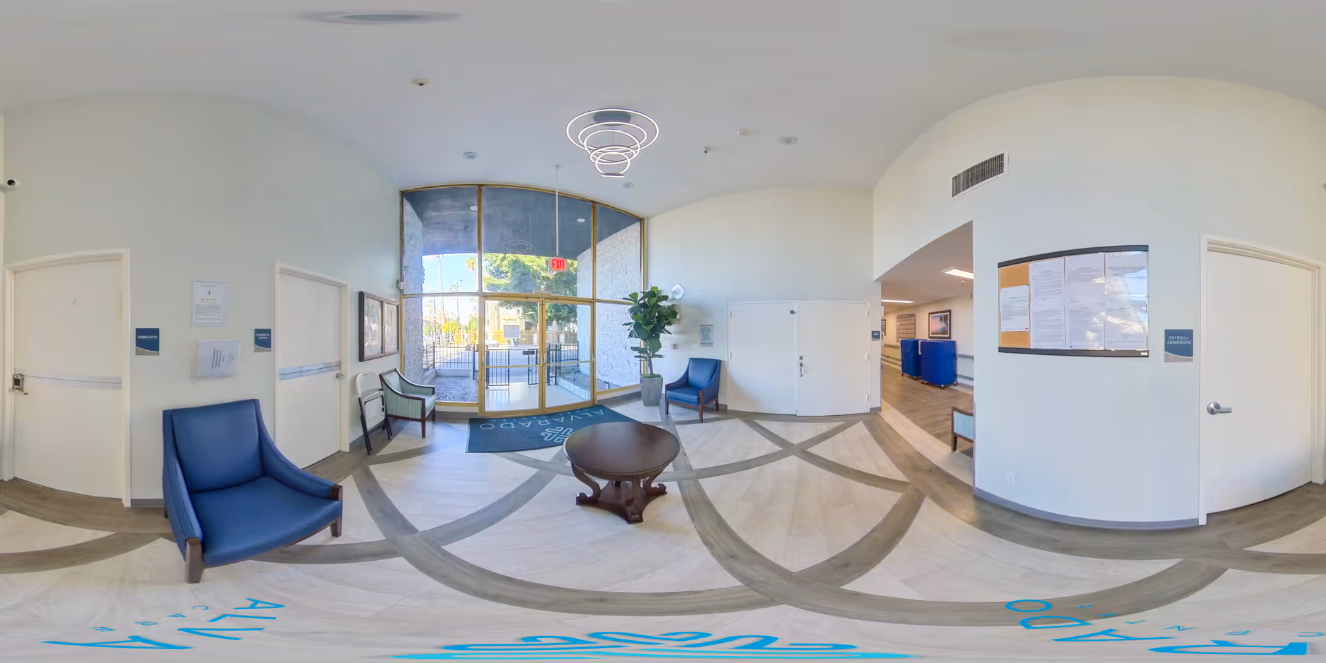 A bright and spacious lobby area of Alvarado Care Center with light-colored walls and flooring featuring a circular pattern. There are several blue and green chairs arranged around a small round wooden table. Large glass doors provide a view of the outdoor area with trees and a gated entrance. A modern circular light fixture hangs from the ceiling, and a bulletin board is mounted on the wall near a hallway entrance.