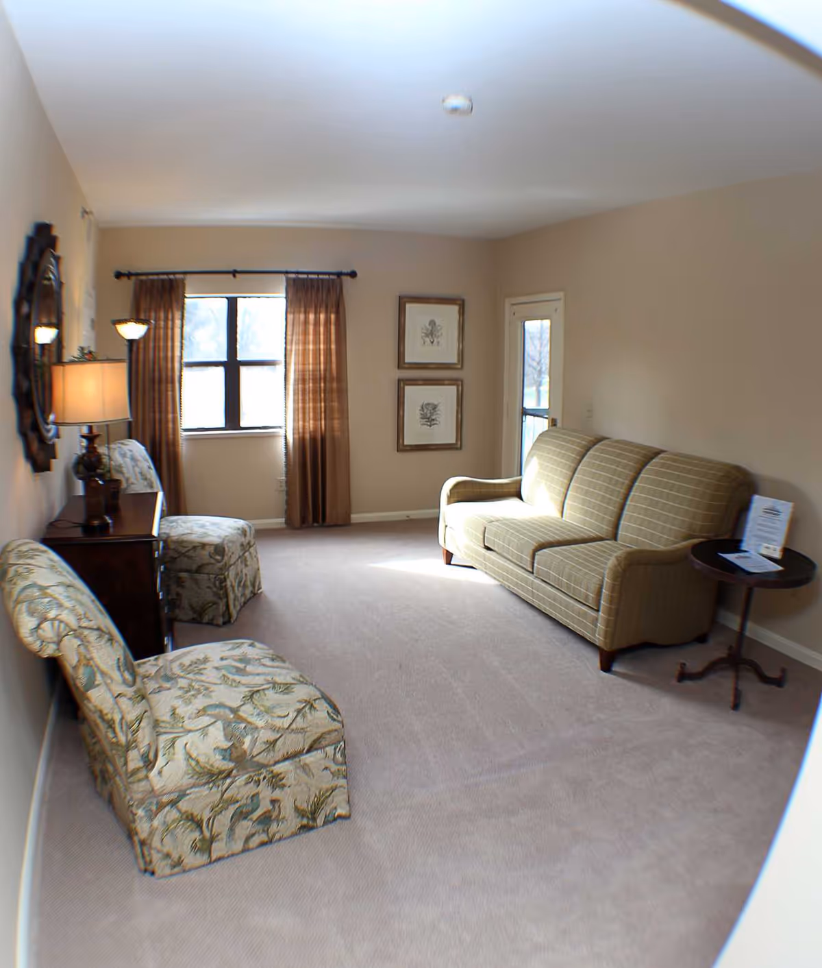 A living room with beige walls and carpet, featuring a green plaid sofa, two floral patterned chairs, a wooden side table with a lamp, a round side table with papers on it, two framed botanical prints on the wall, and a window with brown curtains letting in natural light.