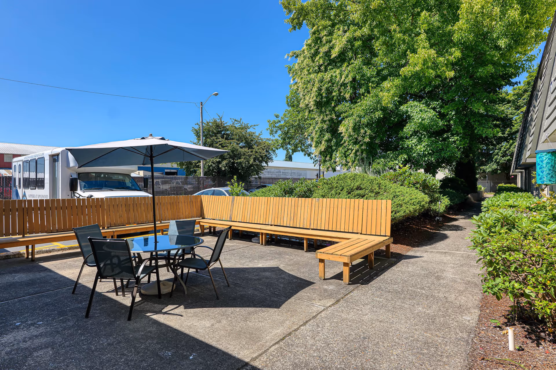 Outdoor patio area with a round glass table, four black chairs, and a large umbrella providing shade. There is a wooden L-shaped bench along the fence and lush green trees and bushes surrounding the area. A white shuttle bus and some parked cars are visible in the background under a clear blue sky.