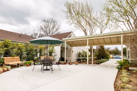 Outdoor patio area at Lincoln Glen Manor with a round table, four chairs, and a large green umbrella. There is a wooden bench to the left, surrounded by various plants and shrubs. The area is paved with concrete and partially covered by a roofed walkway. Trees and a cloudy sky are visible in the background.