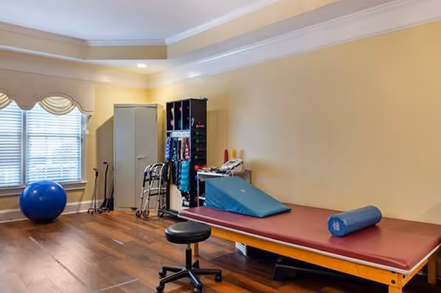 A therapy or exercise room with a padded treatment table, a blue exercise ball, a walker, resistance bands on a rack, and a rolling stool. The room has wooden floors, beige walls, and a window with blinds and valances.