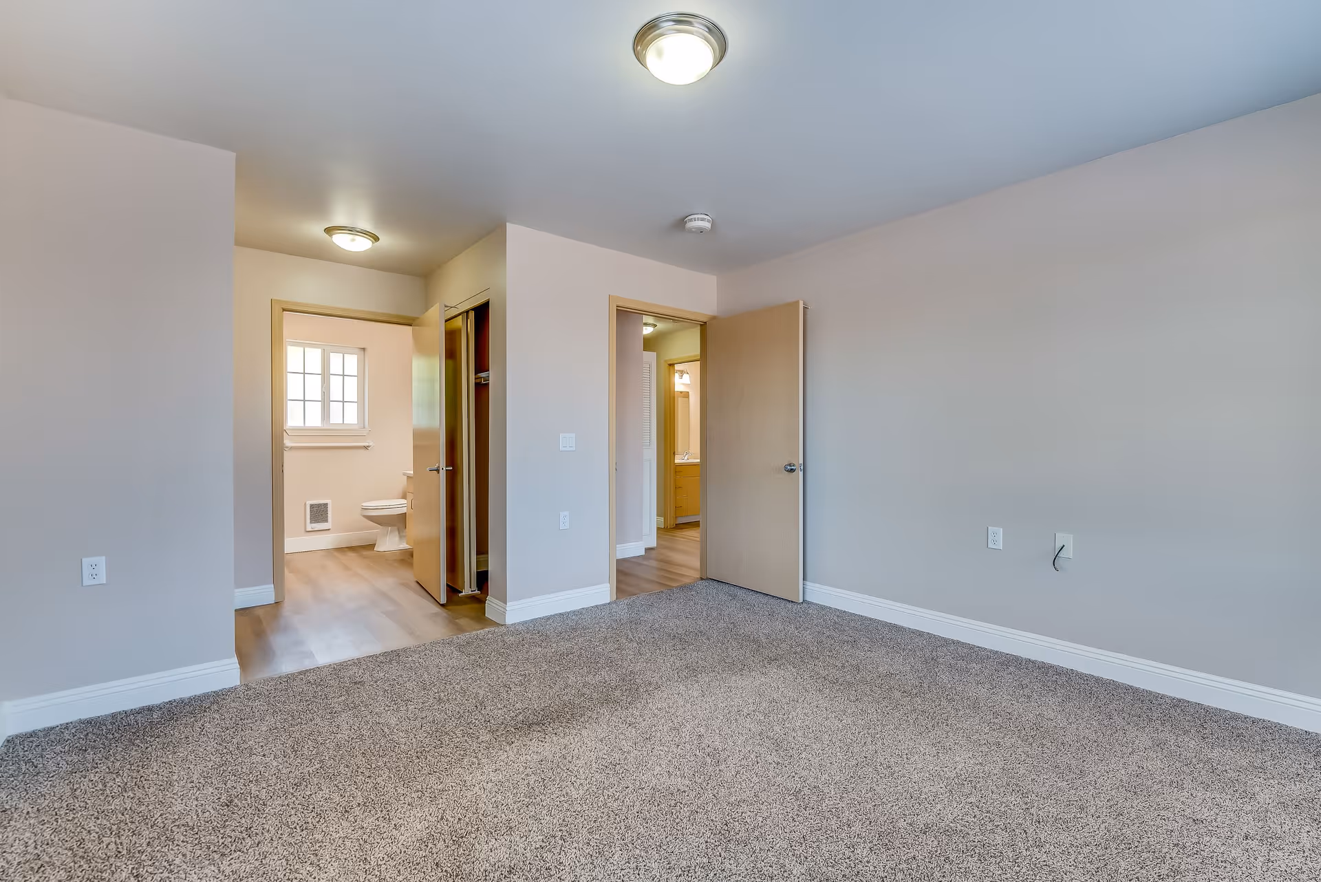 Empty room with beige carpet and light gray walls, featuring two open doorways. One doorway leads to a bathroom with a visible toilet and window, and the other doorway leads to a hallway with a glimpse of a vanity and mirror. The room has ceiling lights and white baseboards.