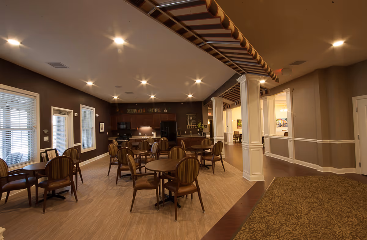 Spacious senior living common room with round dining tables, chairs, and a kitchenette under warm recessed lighting.