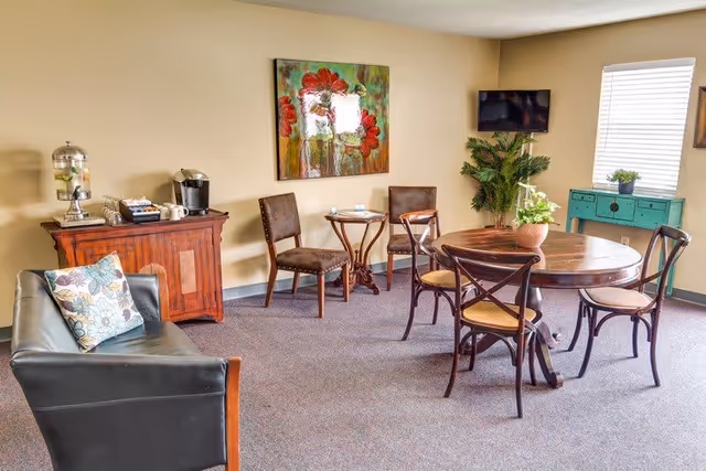 Well-lit common room with a round wooden dining table and chairs, a small sofa, beverage station, wall art and a mounted TV.