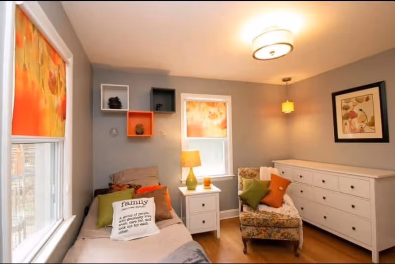 A cozy bedroom with a single bed adorned with multiple pillows including one with the word 'family' printed on it. The room features two windows with orange floral blinds, a white nightstand with a green lamp and a candle, a floral armchair with green and orange pillows, a white dresser with multiple drawers, and wall shelves in white, orange, and gray. The walls are painted gray and decorated with a framed floral artwork.