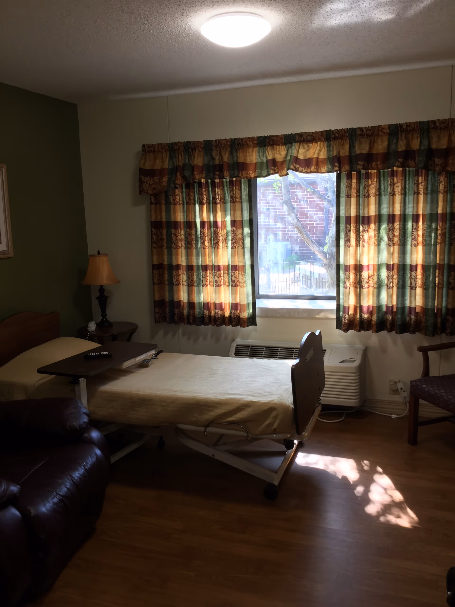 A senior living bedroom with an adjustable bed, recliner, side table and lamp, and a window with patterned curtains.