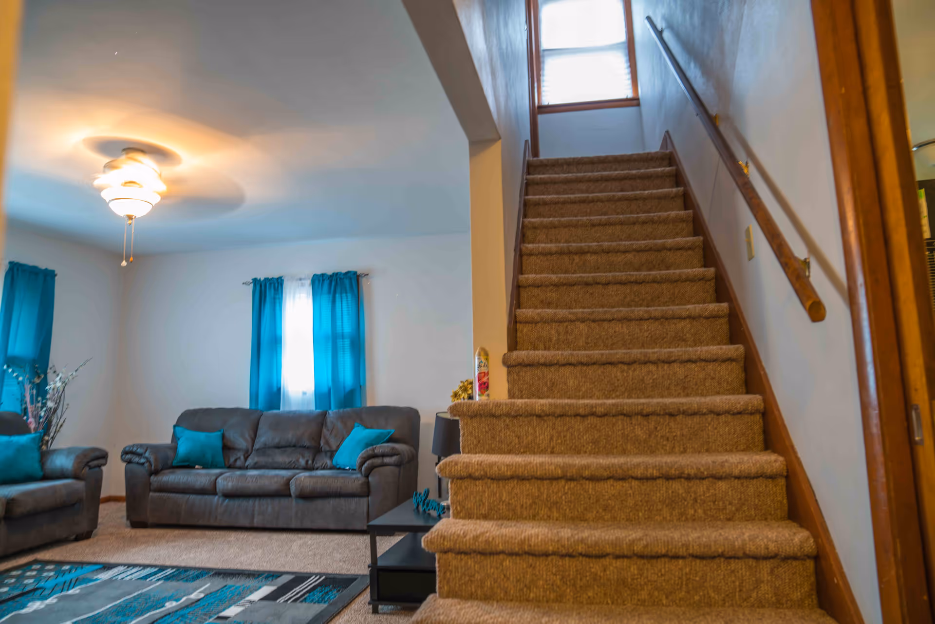 Interior view of a living room area with a carpeted staircase leading upstairs. The living room has a gray sofa and armchair with teal cushions, teal curtains on the windows, a ceiling fan with light, and a patterned area rug in shades of teal, gray, and black.