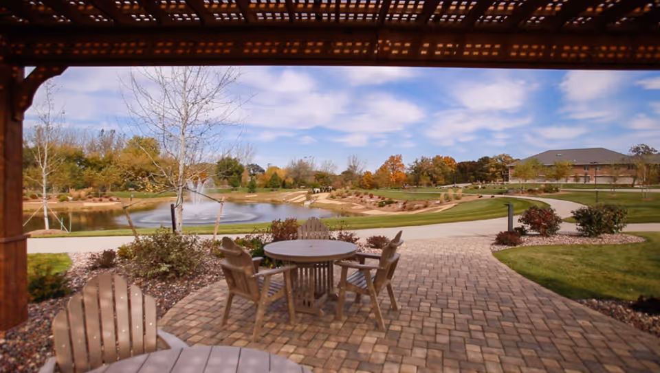 View from under a wooden pergola showing a paved patio area with a round table and four wooden chairs. Beyond the patio is a landscaped garden with a pond featuring a water fountain, walking paths, trees, and a building in the distance under a partly cloudy sky.