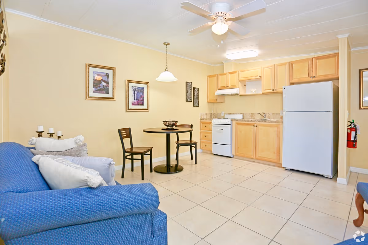 A small living area with a blue couch and white pillows, a round dining table with two chairs, and a compact kitchen featuring light wood cabinets, a white refrigerator, and a white stove. The walls are painted light yellow, and there are framed pictures and decorative items on the walls. The floor is tiled, and a ceiling fan with a light is mounted on the ceiling.