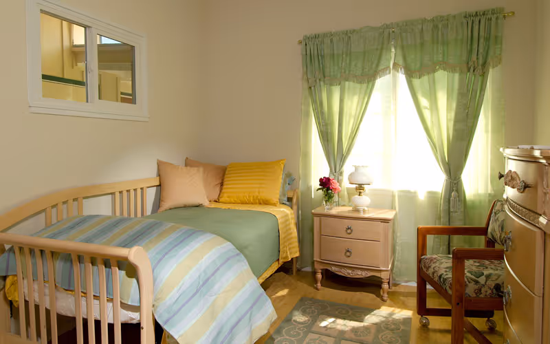 A small, cozy bedroom with a single wooden bed covered with a striped blue and green blanket and yellow pillows. Next to the bed is a wooden nightstand with a white lamp and a vase of flowers. Green curtains cover a window letting in sunlight. There is a wooden chair with floral upholstery and a wooden dresser on the right side of the room.