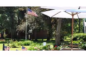 Sunlit outdoor garden area with American flags, patio umbrellas, trees, and seating.