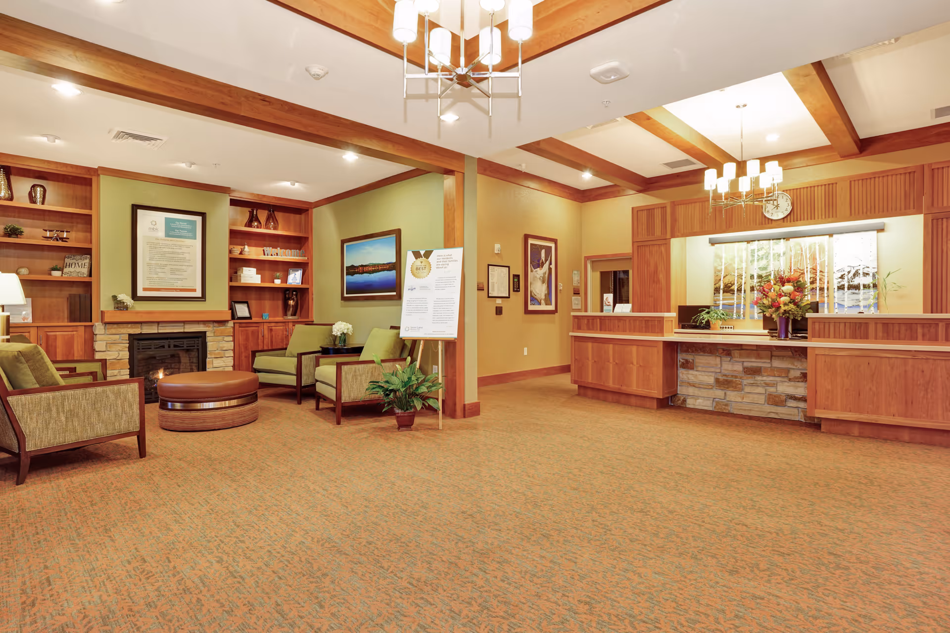 A spacious and warmly lit reception area in a senior living facility featuring a wooden front desk with a stone base, a floral arrangement, and a large window with a nature-themed blind. To the left, there is a cozy seating area with green upholstered chairs, a round ottoman, a fireplace with a stone surround, and built-in wooden shelves displaying decorative items. The room has beige walls, wooden ceiling beams, and carpeted flooring.