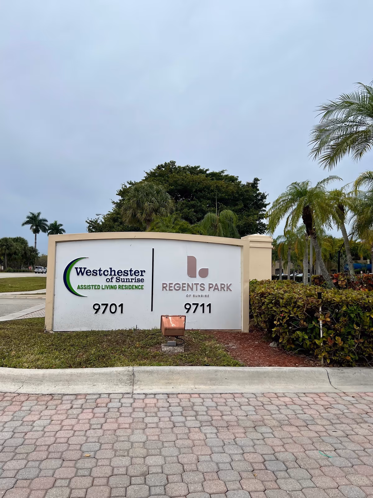 Outdoor view of a large sign at the entrance of two assisted living facilities, Westchester of Sunrise and Regents Park of Sunrise, with their respective addresses 9701 and 9711. The sign is surrounded by palm trees and greenery under a cloudy sky.