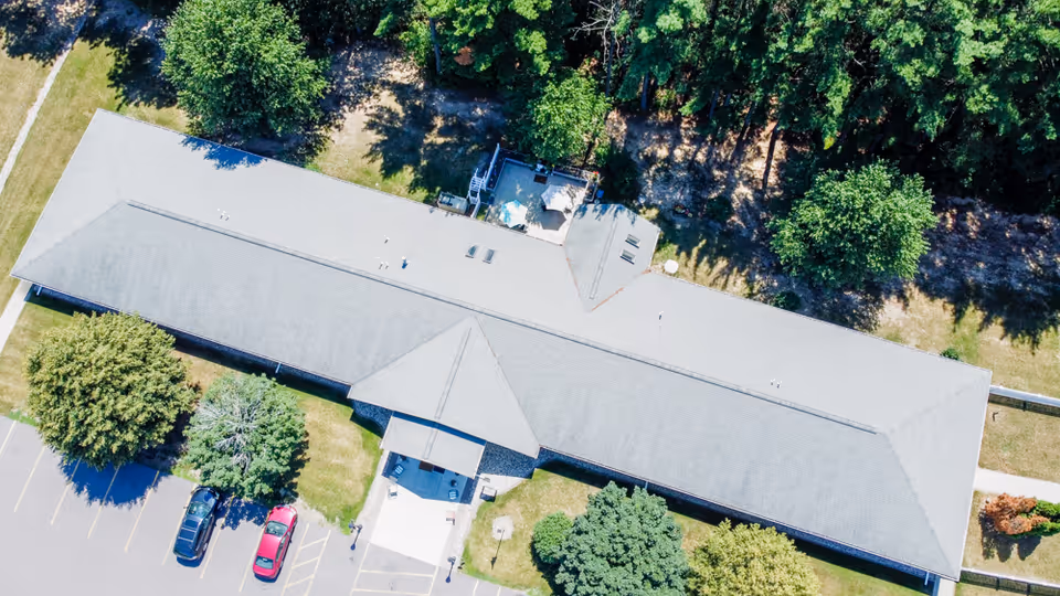 Aerial view of a long, single-story building with a gray roof surrounded by trees and greenery. There is a parking lot with a few cars parked near the entrance, and a small patio area with outdoor furniture is visible behind the building.