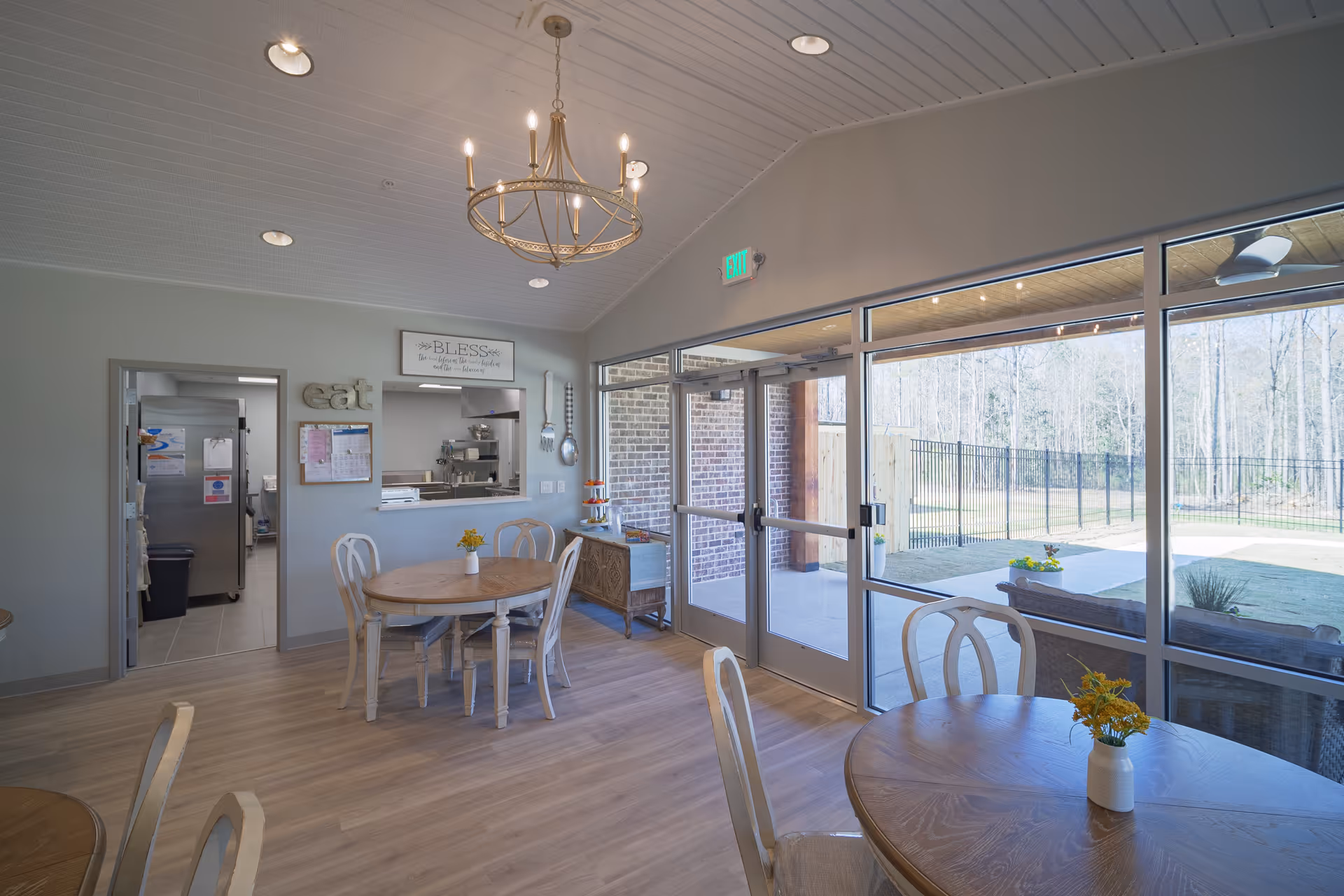 A bright dining area with round wooden tables and white chairs, each table decorated with a small vase of yellow flowers. The room has large glass doors and windows providing a view of an outdoor patio and fenced yard with trees in the background. A serving window opens to a kitchen area, with decorative wall hangings including the word 'eat' and a sign that reads 'BLESS the food before us, the family beside us, and the love between us.' A chandelier hangs from the white paneled ceiling.