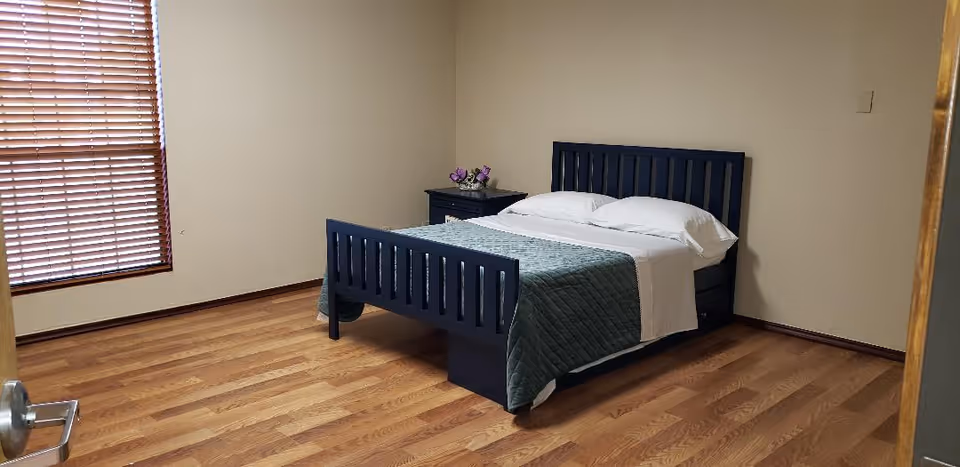 A simple bedroom with a wooden floor, a dark blue bed frame with a neatly made bed featuring white pillows and sheets, and a green quilt. There is a small dark blue nightstand with a flower arrangement on it next to the bed. A window with closed wooden blinds is on the left wall.