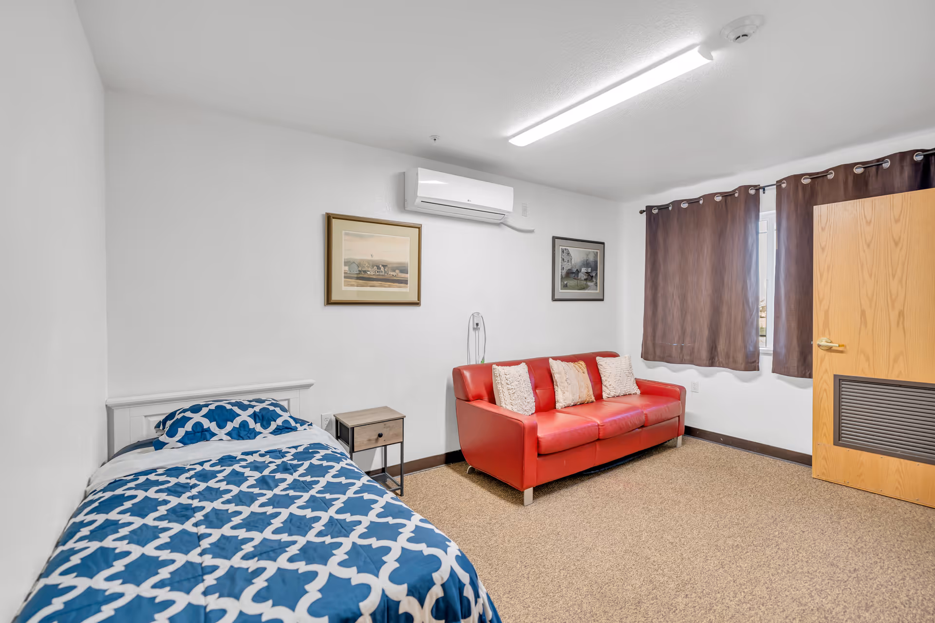 A simple bedroom with a single bed covered in a blue and white patterned comforter, a small wooden nightstand, a red sofa with three white pillows, two framed pictures on the wall, a window with brown curtains, and a wooden door with a vent panel. The room has white walls and a beige carpeted floor.