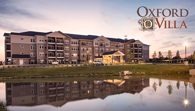 Exterior view of a large three-story senior living apartment building named Oxford Villa, reflected in a calm pond in the foreground during dusk with lights on inside the building.