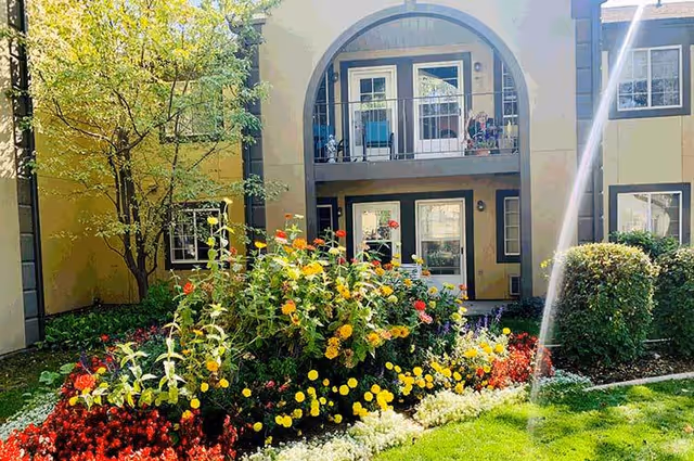 A vibrant garden with colorful flowers and green shrubs in front of a two-story building with an arched balcony and multiple windows and doors.