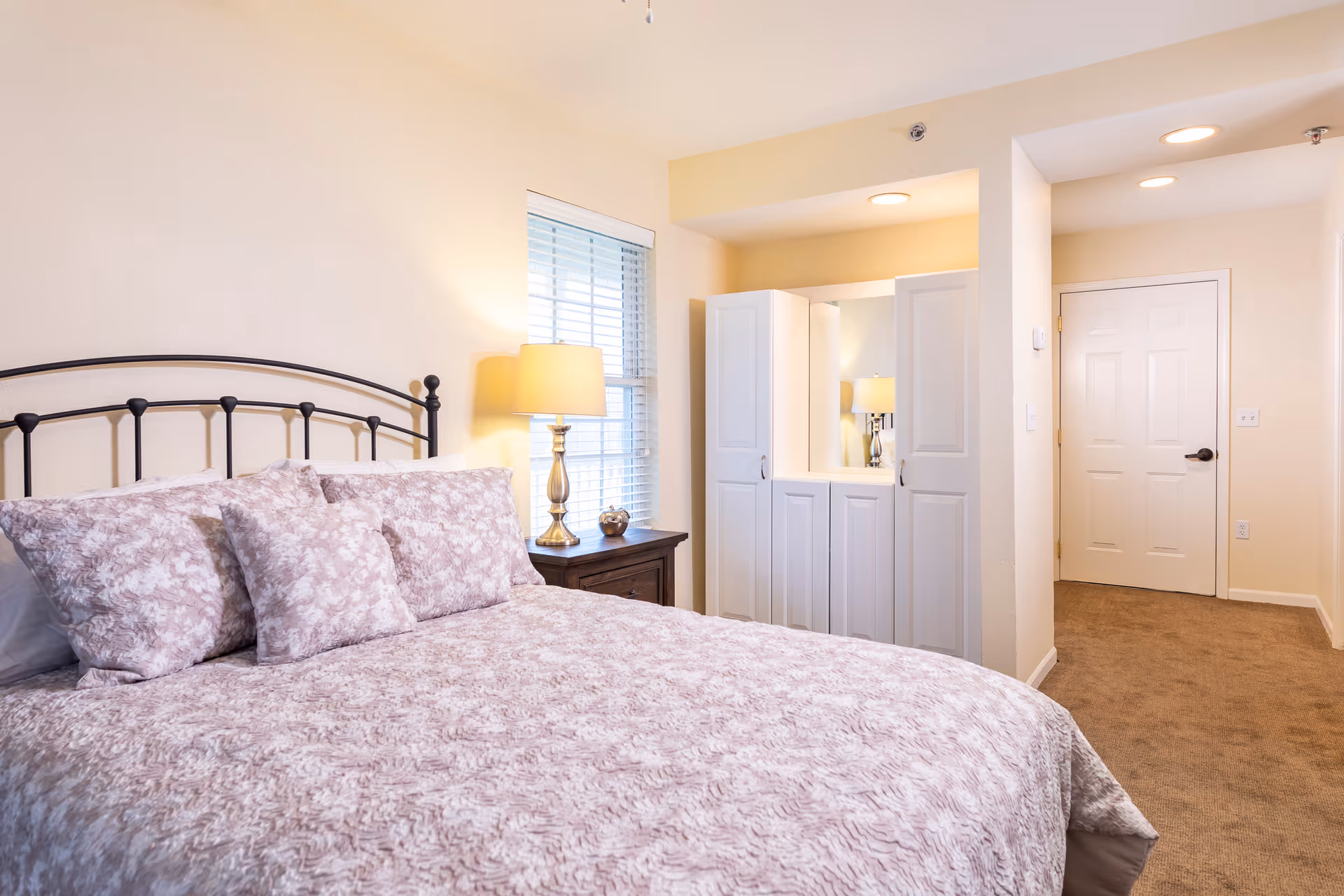 A bedroom with a bed featuring a metal headboard and floral-patterned bedding. Next to the bed is a wooden nightstand with a lamp and a small decorative item. A window with blinds is behind the nightstand. Across from the bed is a built-in vanity with a mirror and cabinets, and a hallway leading to a closed door.