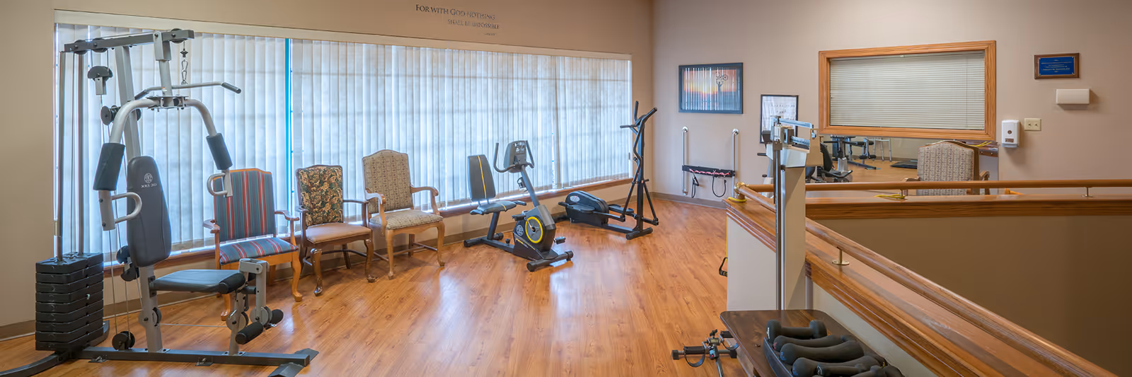 A small exercise room with wooden flooring featuring various fitness equipment including a multi-gym machine, a stationary bike, an elliptical trainer, and a set of dumbbells on a table. There are four upholstered chairs lined up against a large window with vertical blinds. The walls are beige with framed pictures and a large mirror. A motivational quote is visible above the window.
