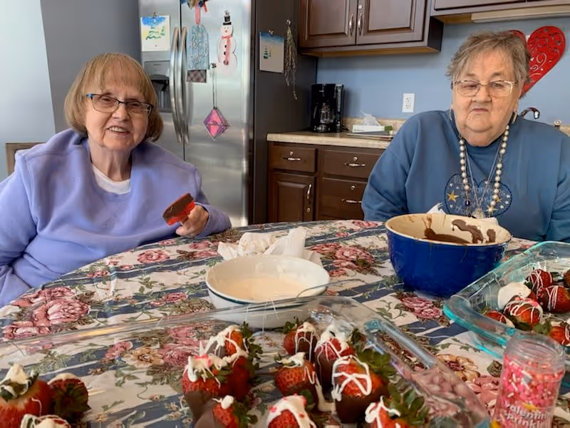 Two elderly women sitting at a kitchen table covered with a floral tablecloth. They are surrounded by bowls and trays filled with chocolate-covered strawberries and other dessert ingredients. One woman is holding a chocolate-covered strawberry, and the kitchen cabinets and refrigerator are visible in the background.