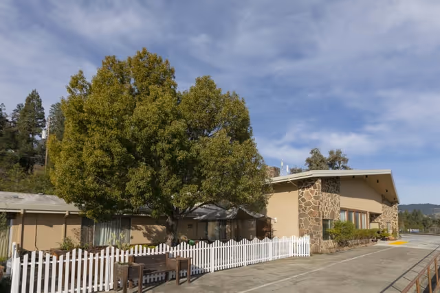 Exterior view of Cloverdale Healthcare Center showing a single-story building with stone and beige walls, large windows, a white picket fence, a large green tree, and a clear blue sky with some clouds.