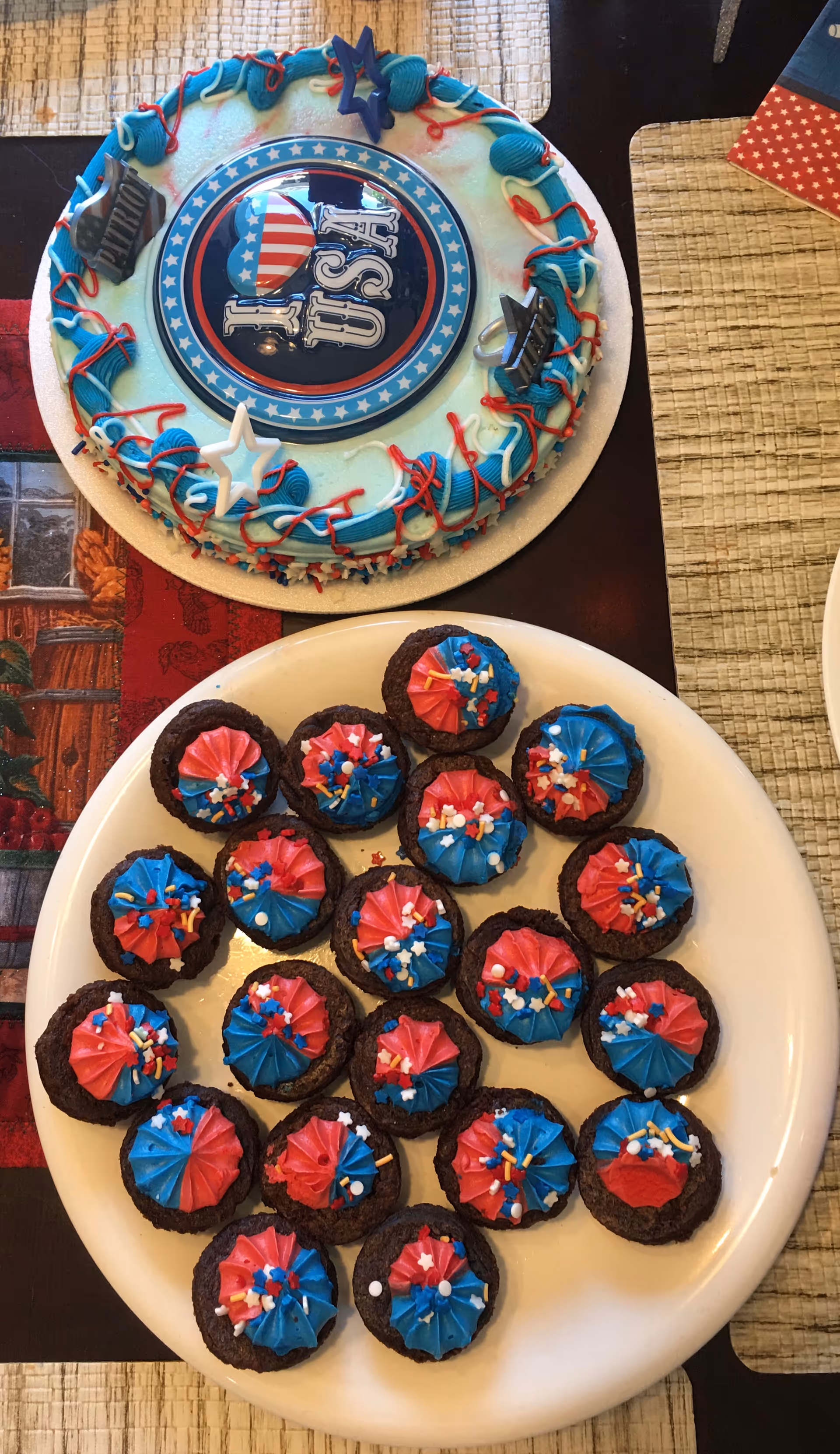 A round cake decorated with blue and red icing and patriotic decorations including a circular topper that says 'I ♥ USA' with an American flag design. Below the cake is a white plate filled with chocolate cookies topped with red and blue icing and colorful sprinkles. The items are placed on a table with a woven placemat and a red and black table runner.