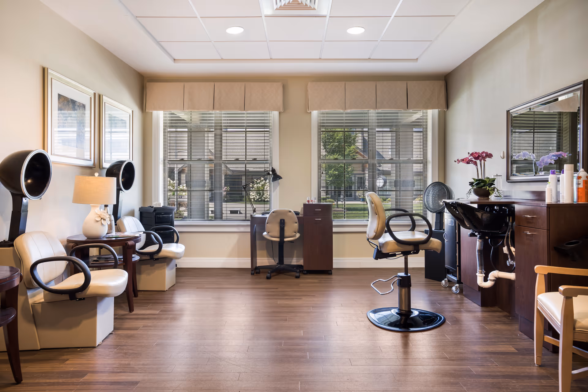 A well-lit hair salon area inside a senior living facility with two hair drying chairs on the left, a styling chair in the center, and a wash basin with hair care products on the right. Large windows with blinds allow natural light to fill the room, and the floor is wooden.