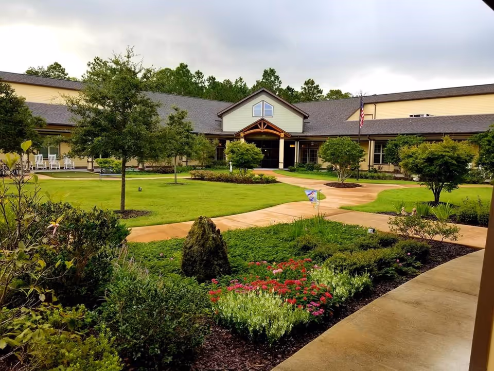 View of a senior living facility building with a well-maintained garden and green lawn in the foreground. The building has a peaked roof and an entrance with a covered porch. There are several small trees, shrubs, and colorful flowers along a paved walkway leading to the entrance. An American flag is visible near the entrance.